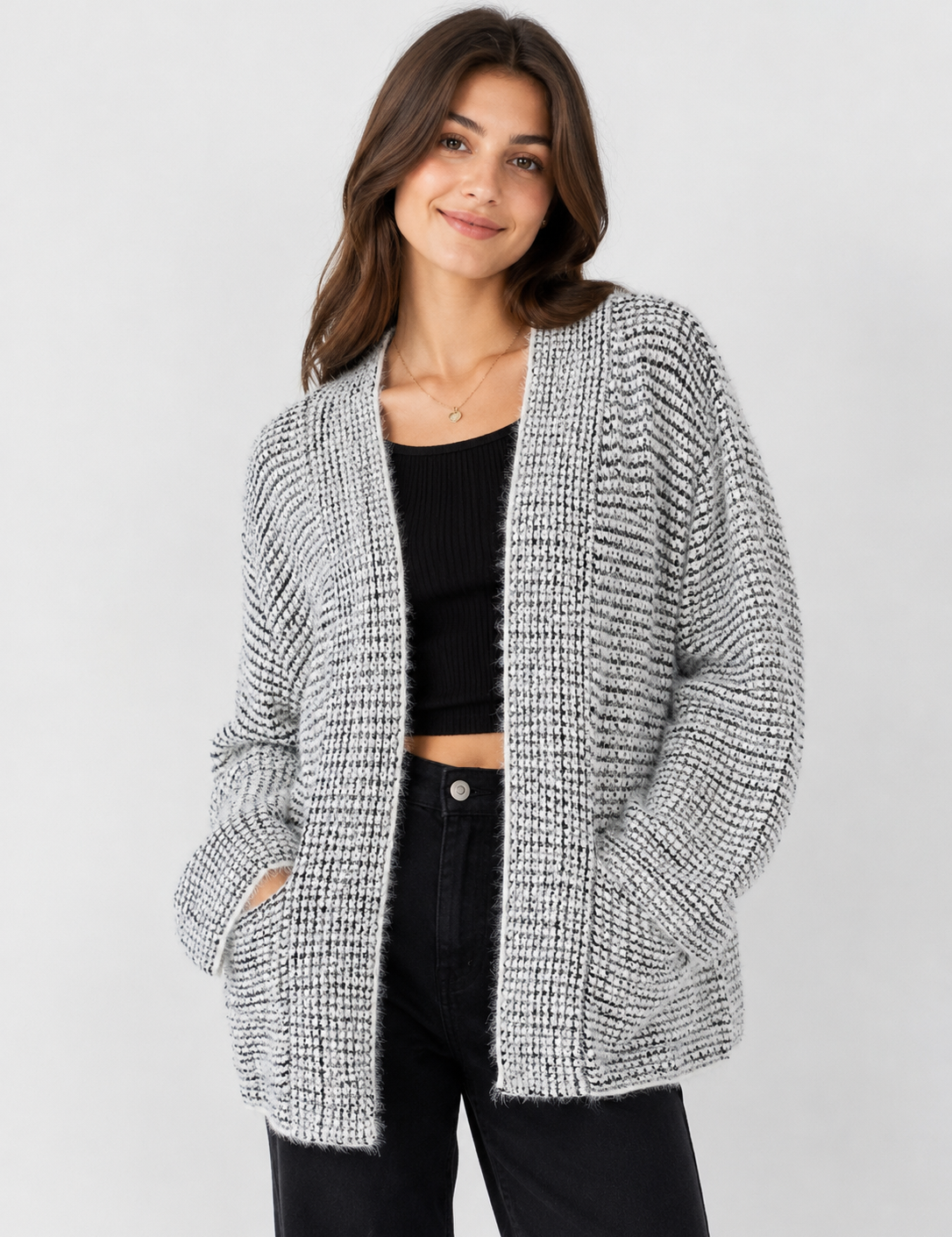 A woman modeling a chunky knit open-front cardigan in a black and white textured pattern, worn over a black crop top and black pants against a light gray studio background.