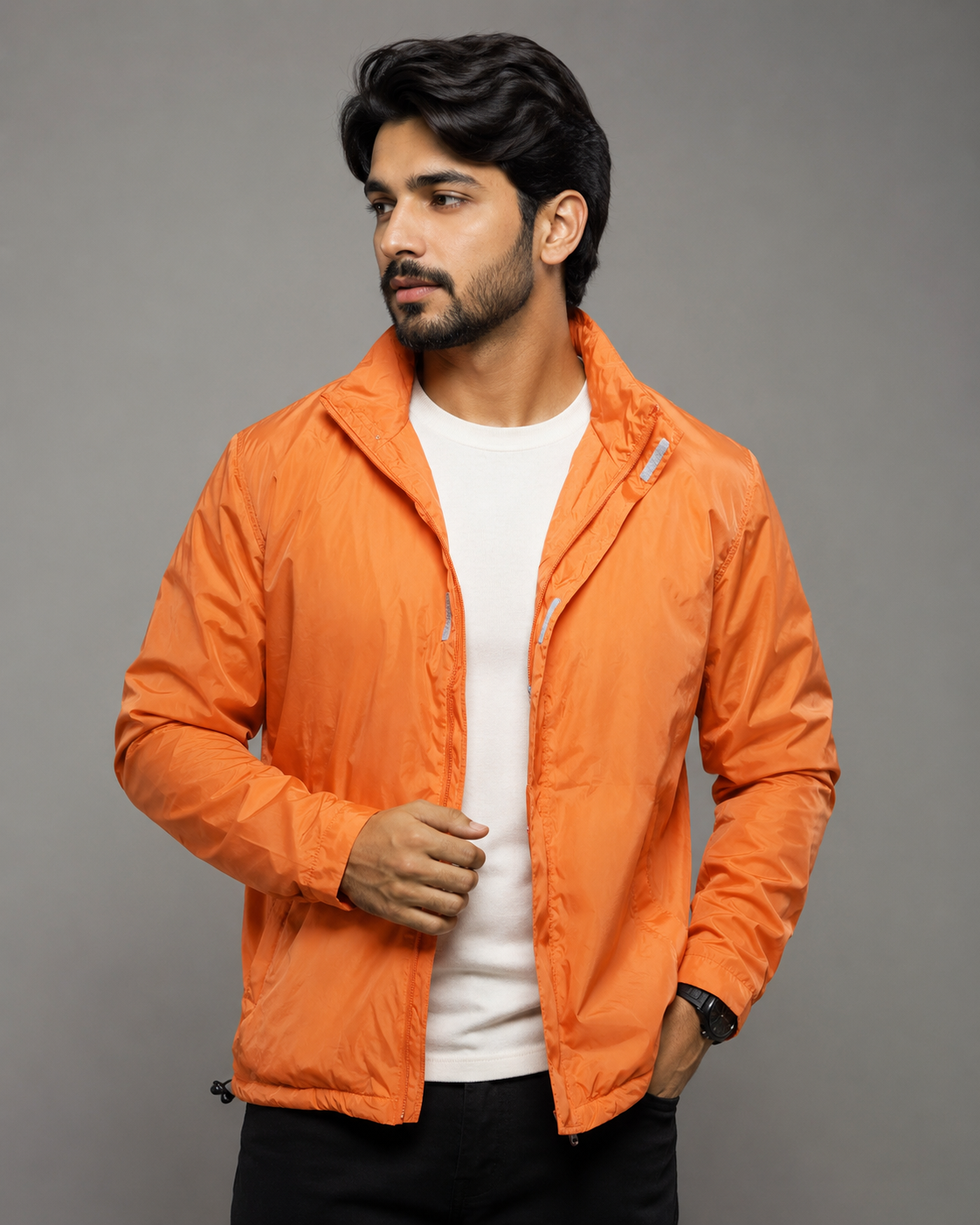 A man wearing a bright orange lightweight zip-up jacket over a white t-shirt and black pants, standing against a solid gray studio background.