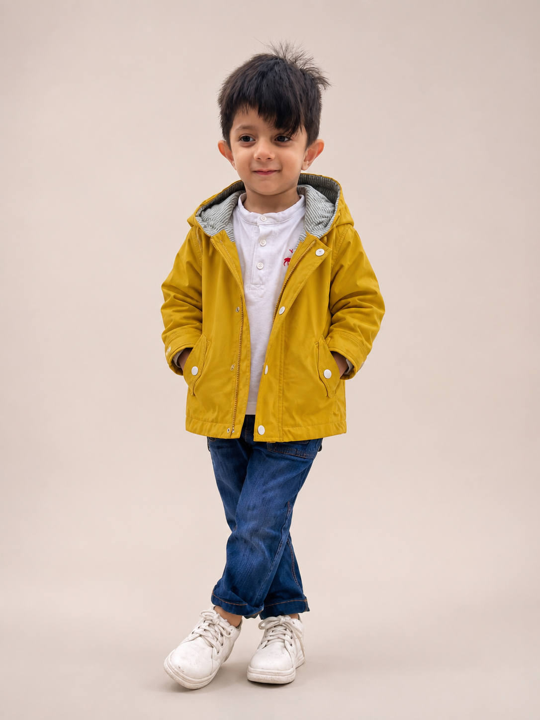 A young boy modeling a mustard yellow hooded jacket over a white shirt and blue jeans, standing against a solid beige studio background.