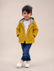 A young boy modeling a mustard yellow hooded jacket over a white shirt and blue jeans, standing against a solid beige studio background.