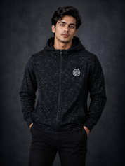 A man wearing a F&F black and white heathered zip-up hoodie with a circular white logo on the left chest, standing against a dark textured studio background.