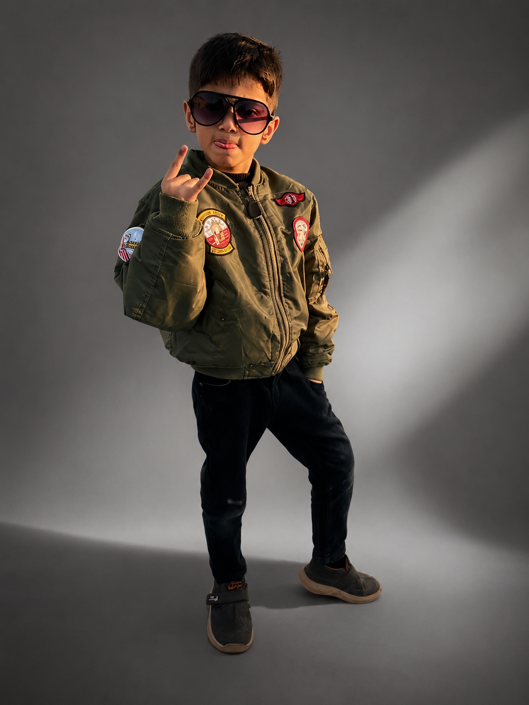 A young boy modeling an olive green bomber jacket with colorful patches, black pants, and sunglasses, posing against a gray studio background with dramatic lighting.