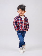 A young boy modeling a red, blue, and white plaid zip-up jacket over a white t-shirt, paired with blue jeans and white sneakers, standing against a light gray studio background.