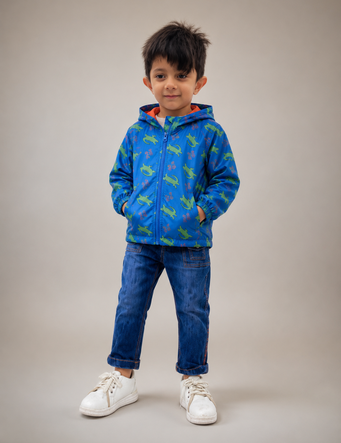 A young boy wearing a blue zip-up hoodie with a green dinosaur pattern, blue jeans, and white sneakers, standing against a solid beige studio background.