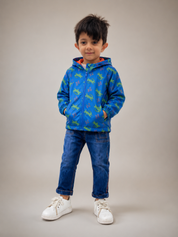 A young boy wearing a blue zip-up hoodie with a green dinosaur pattern, blue jeans, and white sneakers, standing against a solid beige studio background.